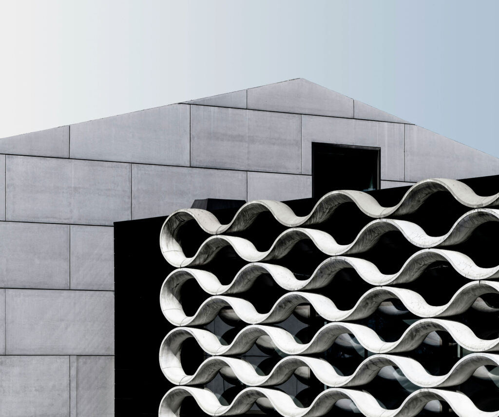Ein modernes Gebäude mit einer Fassade aus überlappenden, gewellten Betonplatten vor einer einfachen grauen, geometrischen Struktur. Das gewellte Muster erzeugt Schatten und Tiefe und bildet einen Kontrast zum flachen, minimalistischen Hintergrund. Der Himmel darüber ist klar und hellblau.